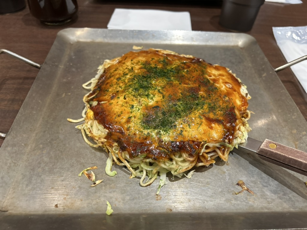 Okonomiyaki de Hiroshima sur une plaque teppan rectangulaire, les couches de nouilles et de chou visibles sur les bords, avec une spatule métallique posée sur le côté