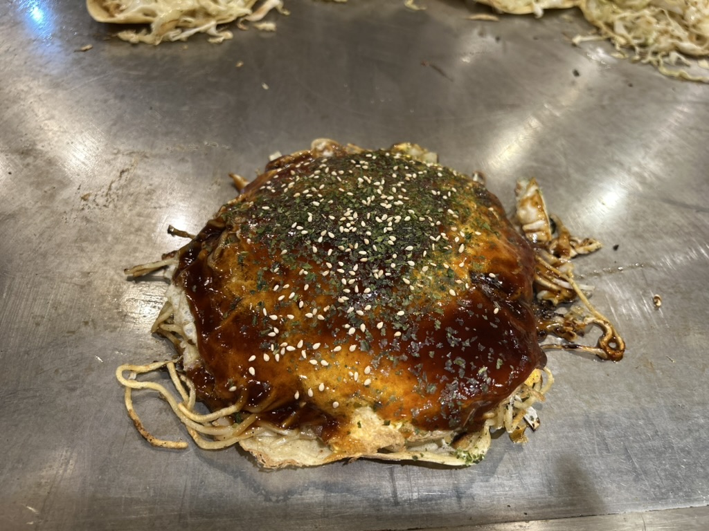 Okonomiyaki de Hiroshima en train de cuire sur une grande plaque de fer, nappé de sauce, d'aonori et de sésame